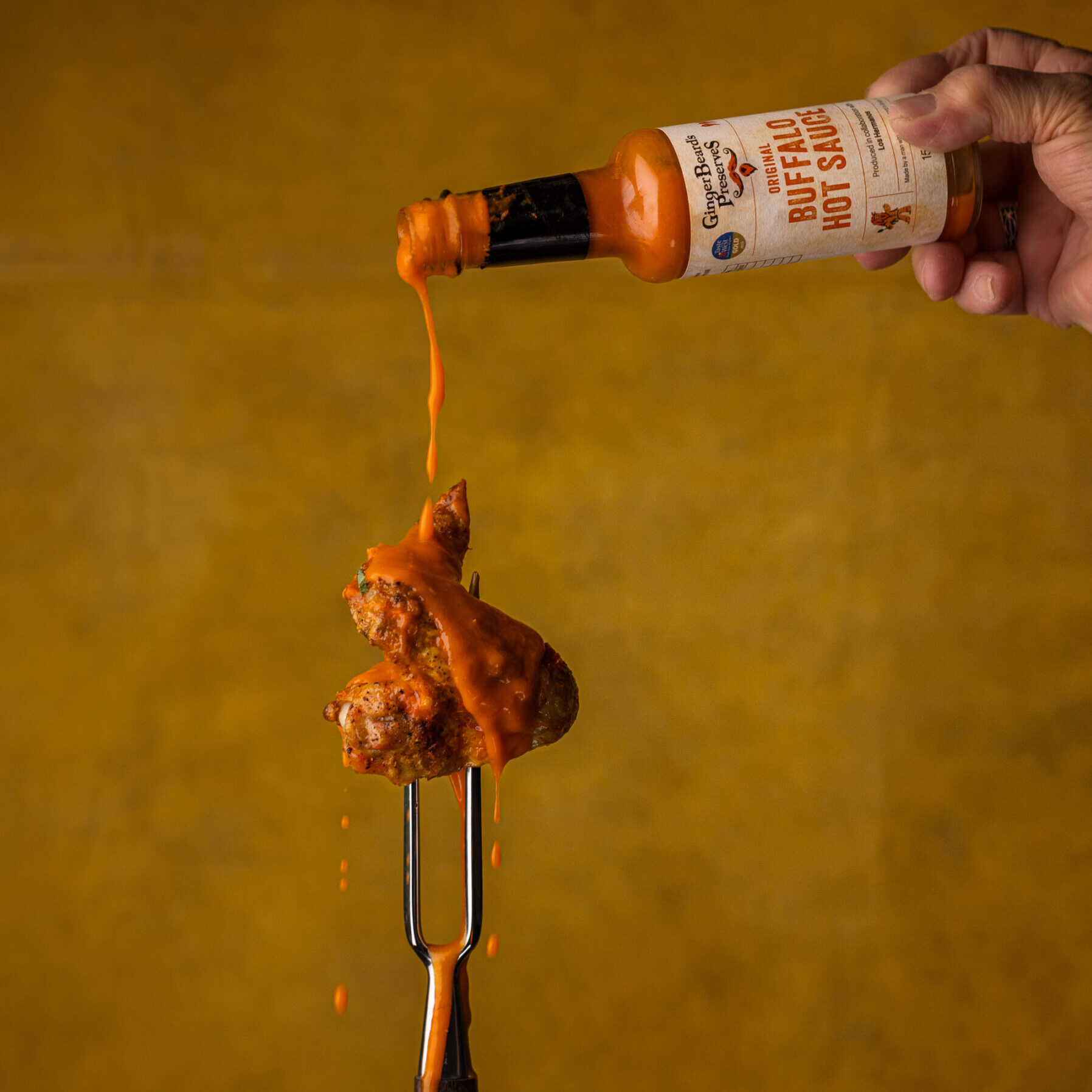 A chicken wing on a fork being drizzled with GingerBeard's Original Buffalo Hot Sauce